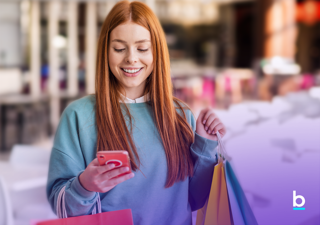 Foto colorida de mulher usando o celular para buscar ofertas na Black Friday. Ela está com várias sacolas de compras nos braços.
