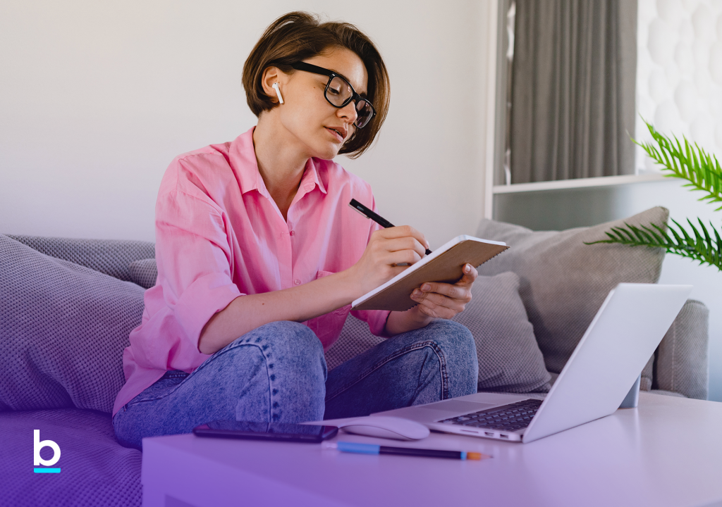 Mulher sentada no sofá com computador na mesa e escrevendo em um bloco seu planejamento financeiro para melhorar seu perfil de crédito.